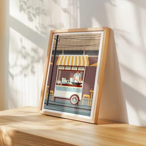 May include: A framed print of a street food cart with a yellow and white striped awning. The illustration depicts a person cooking, with text above the cart. The print is in a light wood frame.