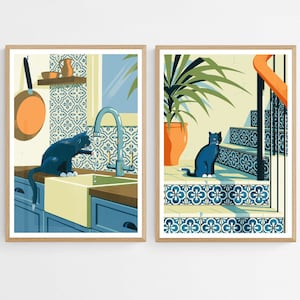 Op de afbeelding: Twee ingelijste illustraties van katten in een keuken en op een trap. De keukenillustratie toont een blauwe kat die drinkt uit een gootsteen met blauw-witte betegelde tegels. De trapillustratie toont een blauwe kat die op een trap zit met blauw-witte betegelde tegels.