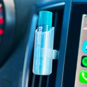 May include: A clear, 3D-printed car vent clip holding a blue bottle with white text. The clip is attached to a car vent, with a car infotainment screen visible in the background.