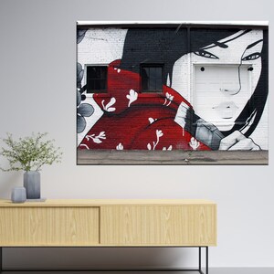May include: A black and white mural on a brick wall depicts a woman with long black hair wearing a red kimono with white floral patterns. The woman's face is partially obscured by the kimono, and she is looking to the side. The mural is painted in a realistic style.