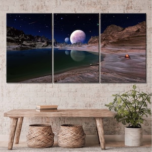 Op de afbeelding: Een driedelig canvasprint met een landschap met een meer, bergen en een grote maan aan de nachtelijke hemel. De maan is lichtroze en paars van kleur met een kleinere maan op de achtergrond. Het meer is donkergroen en de bergen zijn bruin en oranje.