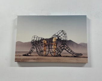 May include: A black metal sculpture of two figures sitting in a desert landscape. The figures are made of wire mesh and are holding each other. The sculpture is a powerful representation of human connection and vulnerability.