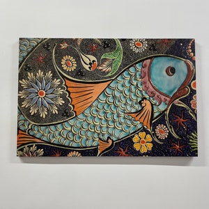 May include: A colorful ceramic tile featuring a blue fish with orange fins and scales. The fish is surrounded by a floral pattern in shades of blue, orange, yellow, and green.