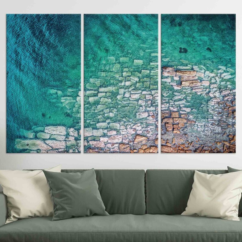 Op de afbeelding: Een driedelige abstracte kunstprint met een close-up luchtfoto van een rotsachtige kustlijn met turquoise water. Het water is helder en ondiep, waardoor de rotsen en het zand eronder zichtbaar zijn. De rotsen zijn een mix van lichte en donkere kleuren, wat een getextureerd en interessant patroon cre&euml;ert.