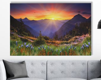 May include: A landscape canvas print depicting a mountain range at sunset. The sky transitions from orange to purple, with the sun's rays bursting through the clouds. Foreground features green grass and blue flowers.