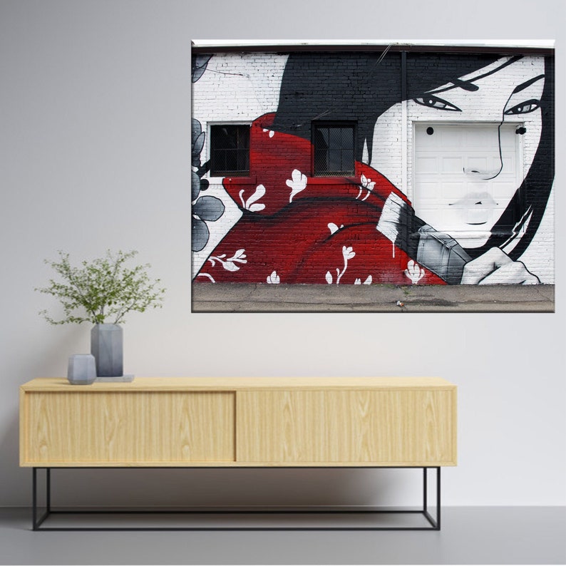 May include: A black and white mural on a brick wall depicts a woman with long black hair wearing a red kimono with white floral patterns. The woman's face is partially obscured by the kimono, and she is looking to the side. The mural is painted in a realistic style.