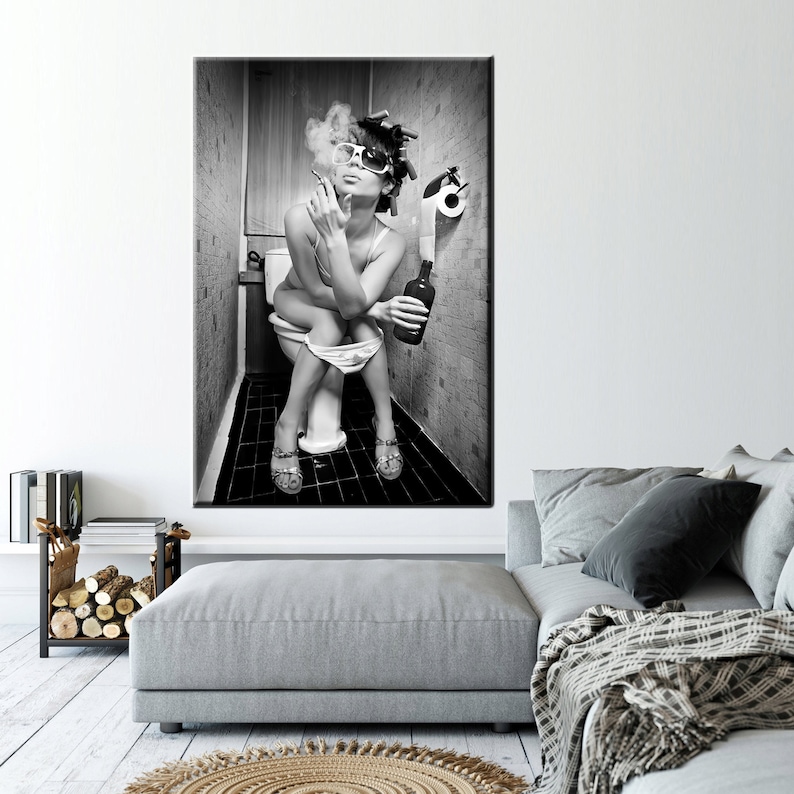 May include: Black and white framed print of a person sitting on a toilet, smoking a cigarette, and holding a bottle. The person is wearing sunglasses and curlers. The bathroom has a roll of toilet paper.