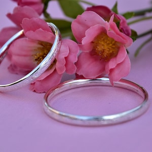May include: Two silver bangle bracelets with a textured design, resting on a pink surface with pink flowers.
