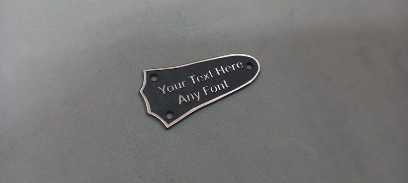 Puede incluir: P&uacute;a de guitarra negra con borde plateado. La p&uacute;a tiene el texto "Your Text Here Any Font" grabado.