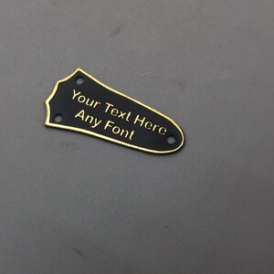 Puede incluir: Golpeador de guitarra negro con ribete dorado y el texto "Your Text Here Any Font" grabado en &eacute;l.