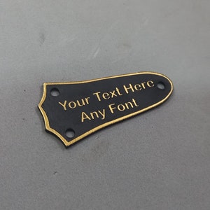 Puede incluir: P&uacute;a de guitarra negra con borde dorado y el texto "Your Text Here Any Font" grabado en oro.