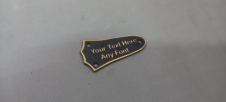 Puede incluir: P&uacute;a de guitarra negra con borde dorado y el texto "Your Text Here Any Font" grabado en oro.