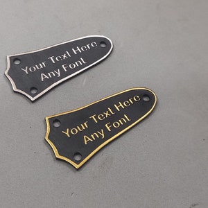 Puede incluir: Dos placas de protecci&oacute;n de guitarra negras con ribetes dorados y plateados. Las placas de protecci&oacute;n tienen agujeros para tornillos de montaje y el texto "Your Text Here Any Font" est&aacute; grabado en cada una.