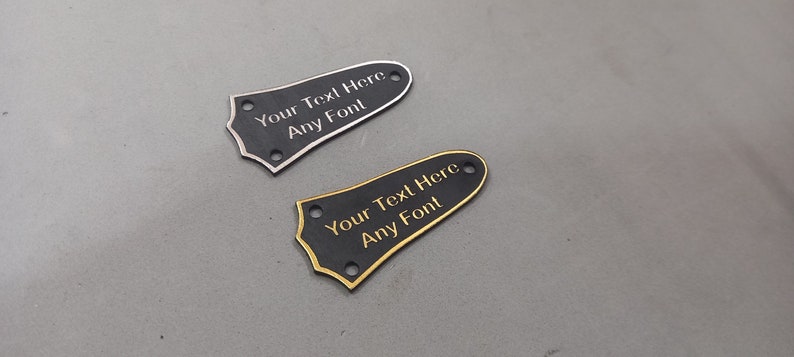 Puede incluir: Dos placas de protecci&oacute;n de guitarra negras con ribetes dorados y plateados. Las placas de protecci&oacute;n tienen agujeros para tornillos de montaje y el texto "Your Text Here Any Font" est&aacute; grabado en cada una.