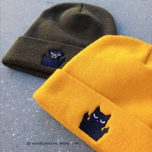 Könnte beinhalten: Zwei Strickmützen mit schwarzen gestickten Katzen. Eine Mütze ist olivgrün und die andere ist gelb. Die Katze auf der gelben Mütze zeigt den Mittelfinger.