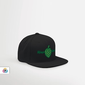 May include: Black baseball cap with a flat brim. The cap features a green embroidered hop design and the words "Hop Time" in a script font. The cap is a solid black color.