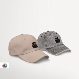 May include: Two baseball caps, one beige and one grey, with embroidered images of a dog and a cat. The dog is black with brown ears and is wearing a red collar. The cat is black and is giving the middle finger.