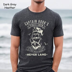 May include: Dark grey heather t-shirt featuring a graphic of a pirate ship with the text "CAPTAIN HOOK'S PIRATE CREW" and "THE JOLLY ROGER + NEVER LAND+". The shirt is short-sleeved.