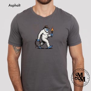 Peut inclure: T-shirt gris avec un dessin d'un y&eacute;ti de dessin anim&eacute; portant un chapeau et tenant une torche et une corde. Le y&eacute;ti est blanc avec des d&eacute;tails bleus. Le texte "Asphalt" est en haut &agrave; gauche, et un logo est en bas &agrave; droite.