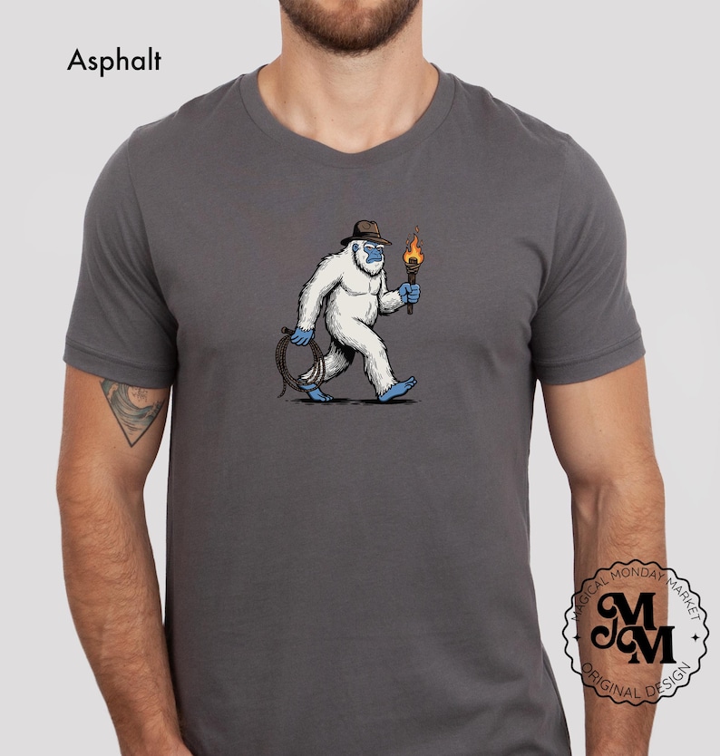 Peut inclure: T-shirt gris avec un dessin d'un y&eacute;ti de dessin anim&eacute; portant un chapeau et tenant une torche et une corde. Le y&eacute;ti est blanc avec des d&eacute;tails bleus. Le texte "Asphalt" est en haut &agrave; gauche, et un logo est en bas &agrave; droite.