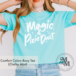 May include: A light blue Comfort Colors boxy tee with the words "Magic & Pixie Dust" in white script. The shirt has short sleeves and is made of a soft material. The shirt is a Chalky Mint color.