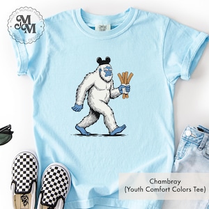 May include: Light blue youth t-shirt with a cartoon yeti wearing a black hat and holding a bundle of churros. The shirt has the text "Chambray (Youth Comfort Colors Tee)" at the bottom. The shirt is next to a pair of black and white checkered Vans shoes.