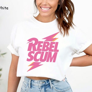 May include: A white short-sleeved t-shirt featuring the text REBEL SCUM in bold pink capital letters. The text is positioned between two pink lightning bolt graphics with orange outlines. The shirt has a crew neckline and cuffed sleeves.