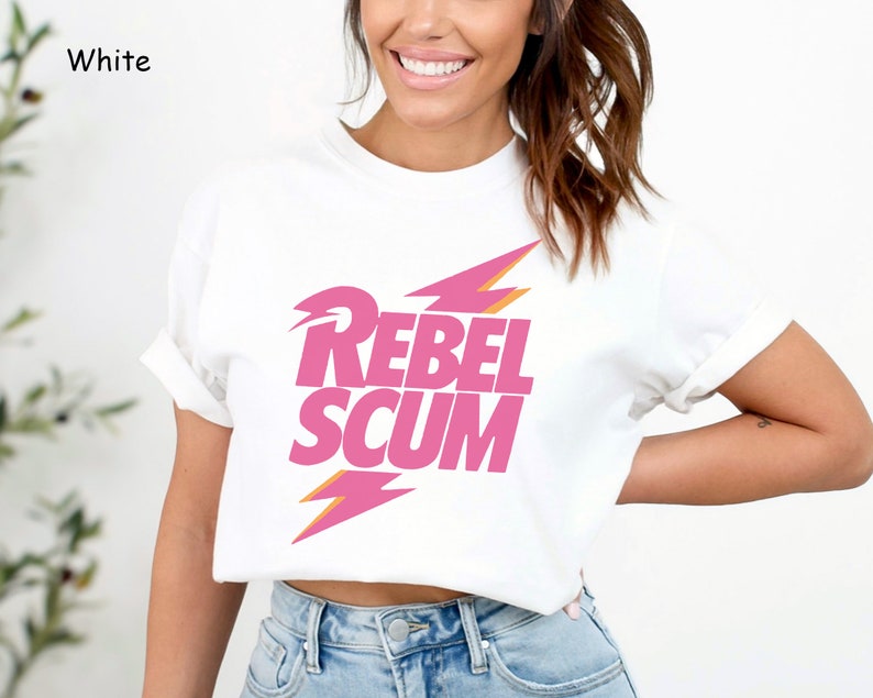May include: A white short-sleeved t-shirt featuring the text REBEL SCUM in bold pink capital letters. The text is positioned between two pink lightning bolt graphics with orange outlines. The shirt has a crew neckline and cuffed sleeves.