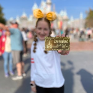 Puede incluir: Una persona joven con una camisa blanca y un lazo amarillo sostiene un boleto dorado de Disneyland con el nombre Natalie Perrin impreso en &eacute;l.