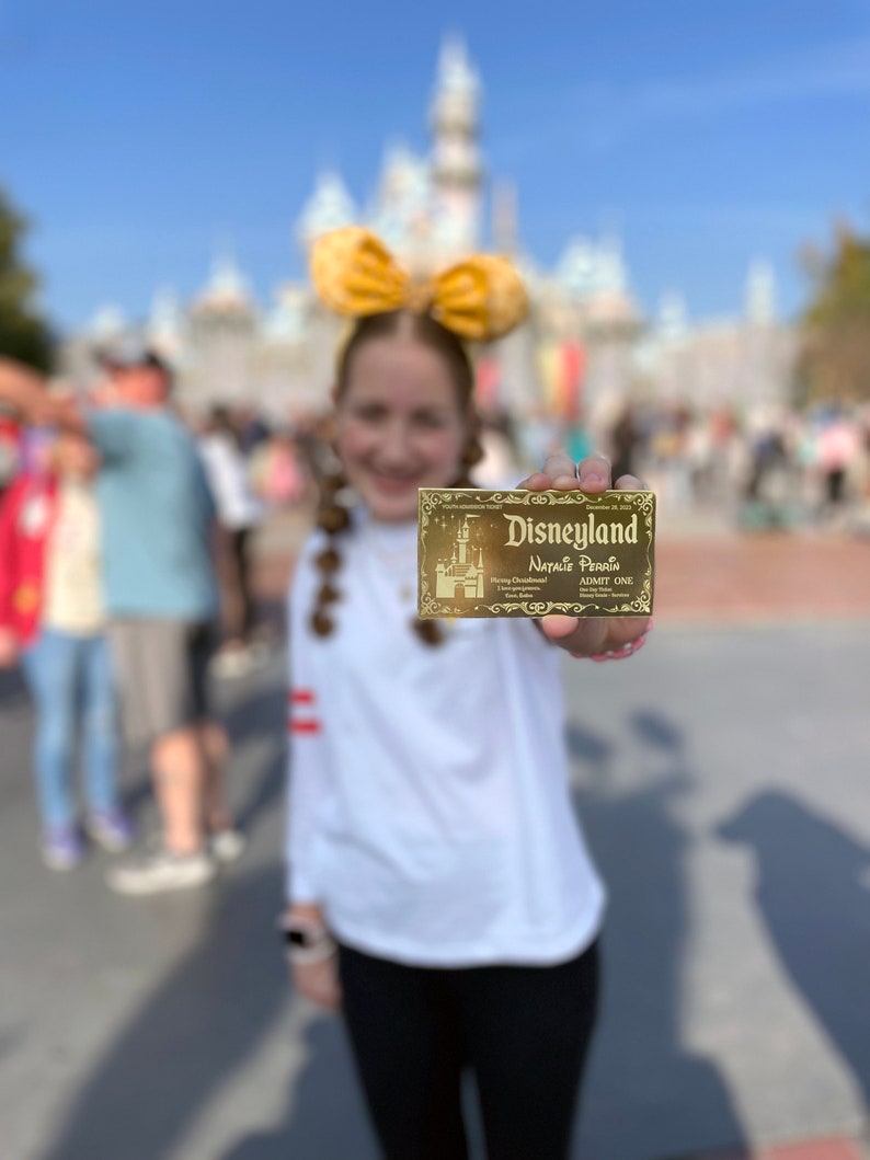 Puede incluir: Una persona joven con una camisa blanca y un lazo amarillo sostiene un boleto dorado de Disneyland con el nombre Natalie Perrin impreso en &eacute;l.