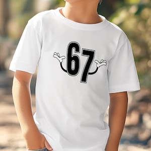 May include: White t-shirt featuring the number 67 in black with cartoon hands. The number is outlined in black and has a playful design. The shirt is short-sleeved and appears to be made of cotton.