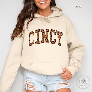 Puede incluir: Una sudadera con capucha de color arena con un estampado de tigre "CINCY". La sudadera tiene una capucha con cordón y un bolsillo canguro.