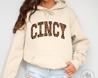 May include: A sand coloured hoodie with a tiger print "CINCY" graphic. The hoodie has a drawstring hood and a kangaroo pocket.