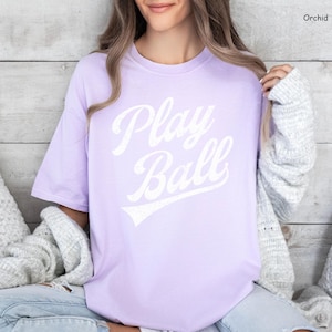 K&ouml;nnte beinhalten: Ein lavendelfarbenes T-Shirt mit dem Text "Play Ball" in wei&szlig;er, vintage-artiger Schrift.