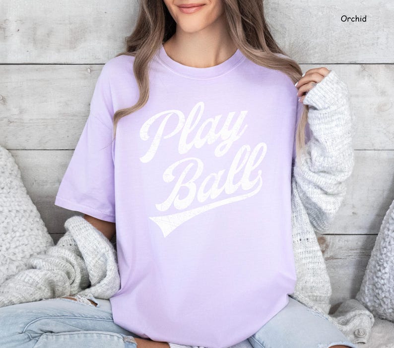 K&ouml;nnte beinhalten: Ein lavendelfarbenes T-Shirt mit dem Text "Play Ball" in wei&szlig;er, vintage-artiger Schrift.