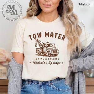 Pu&ograve; includere: T-shirt color crema con grafica marrone di un carro attrezzi e la scritta "TOW MATER". Sotto il camion, il testo recita "TOWING & SALVAGE" e "Radiator Springs". La maglietta ha anche il logo "Magical Monday Market".