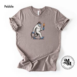 Peut inclure: T-shirt couleur galet avec un dessin d'un y&eacute;ti de dessin anim&eacute; portant un chapeau et tenant une torche et une corde. Le y&eacute;ti marche avec une expression d&eacute;termin&eacute;e. Le t-shirt a des manches retrouss&eacute;es et un col rond. Le logo Magical Monday Market est en bas &agrave; droite.