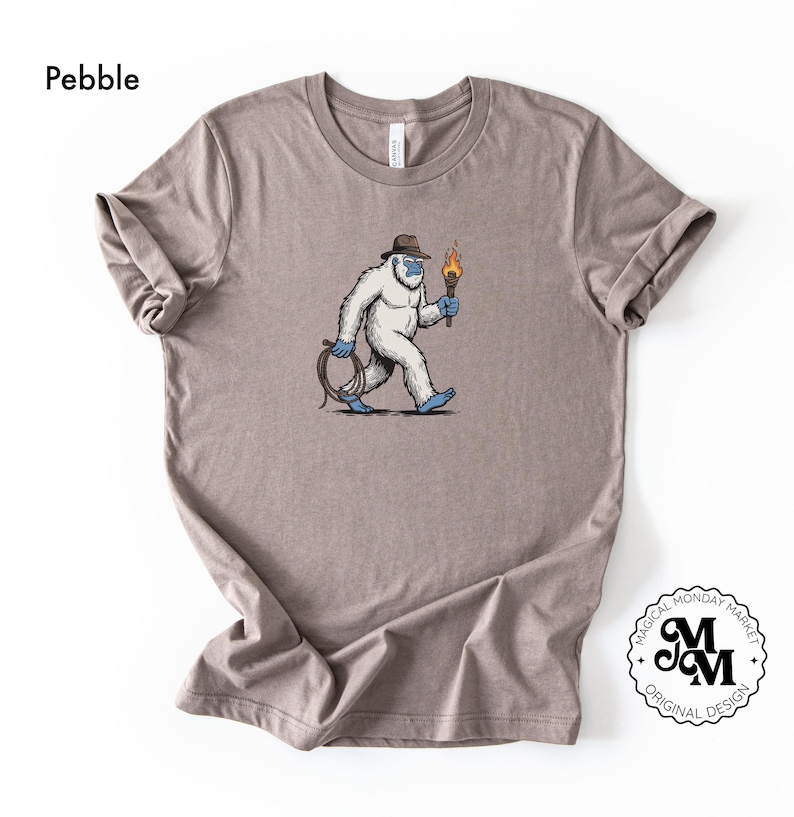 Peut inclure: T-shirt couleur galet avec un dessin d'un y&eacute;ti de dessin anim&eacute; portant un chapeau et tenant une torche et une corde. Le y&eacute;ti marche avec une expression d&eacute;termin&eacute;e. Le t-shirt a des manches retrouss&eacute;es et un col rond. Le logo Magical Monday Market est en bas &agrave; droite.