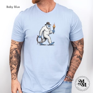 Peut inclure: T-shirt bleu clair avec un dessin de y&eacute;ti de dessin anim&eacute; portant un fedora et tenant une torche et un fouet. Le y&eacute;ti est blanc avec des accents bleus. Le texte "Baby Blue" est au-dessus. Le t-shirt a le logo "Magical Monday Market".