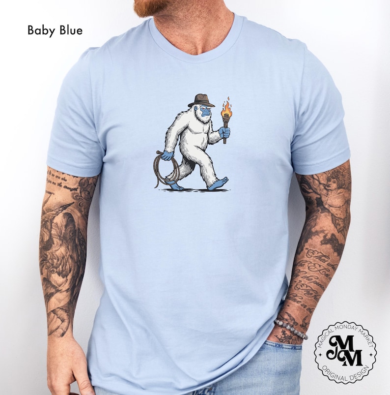 Peut inclure: T-shirt bleu clair avec un dessin de y&eacute;ti de dessin anim&eacute; portant un fedora et tenant une torche et un fouet. Le y&eacute;ti est blanc avec des accents bleus. Le texte "Baby Blue" est au-dessus. Le t-shirt a le logo "Magical Monday Market".