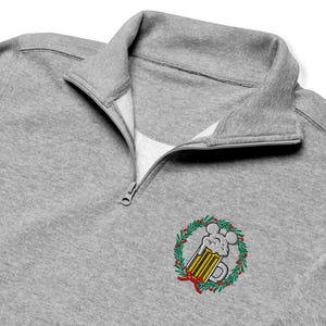 Könnte beinhalten: Ein graumeliertes Sweatshirt mit kurzem Reißverschluss, das eine Stickerei eines Bierkrugs mit schaumigen, mausohrähnlichen Formen zeigt, umgeben von einem festlichen grünen Kranz mit roten Beeren und einer kleinen roten Schleife auf der Brust.