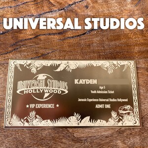 Puede incluir: Un boleto dorado de experiencia VIP de Universal Studios Hollywood con un gr&aacute;fico de dinosaurio. El boleto es para una admisi&oacute;n juvenil y es para Kayden, de 5 a&ntilde;os. El texto "ADMIT ONE" est&aacute; impreso en el boleto.
