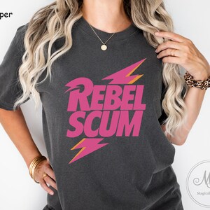 May include: A charcoal gray short-sleeve t-shirt featuring the text REBEL SCUM in bold pink lettering. The design includes two stylized lightning bolt graphics in pink and orange, positioned above and below the text on the chest area.