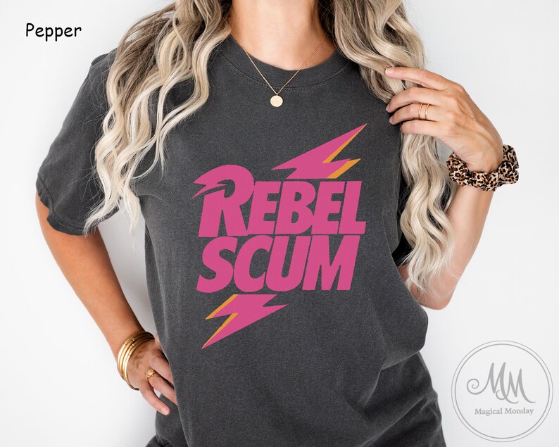 May include: A charcoal gray short-sleeve t-shirt featuring the text REBEL SCUM in bold pink lettering. The design includes two stylized lightning bolt graphics in pink and orange, positioned above and below the text on the chest area.