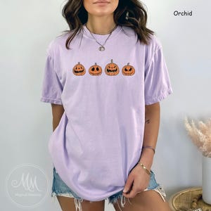 May include: A purple t-shirt featuring a graphic of four orange jack-o'-lantern faces. The t-shirt is a relaxed fit and has a crew neck.