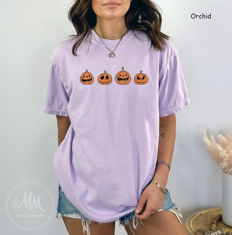 May include: A purple t-shirt featuring a graphic of four orange jack-o'-lantern faces. The t-shirt is a relaxed fit and has a crew neck.