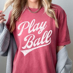 K&ouml;nnte beinhalten: Ein T-Shirt in Rotbraun mit einem wei&szlig;en Vintage-Grafikdruck, auf dem "Play Ball" steht.