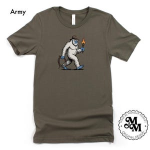 Peut inclure: T-shirt vert arm&eacute;e avec une illustration de dessin anim&eacute; d'un y&eacute;ti blanc portant un chapeau et tenant une torche. Le y&eacute;ti marche et a une corde bleue attach&eacute;e &agrave; sa taille. Le texte "Army" est en haut &agrave; gauche.