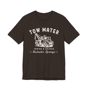 Pu&ograve; includere: T-shirt marrone scuro con una grafica bianca di un carro attrezzi e la scritta "TOW MATER" sopra. Sotto il camion ci sono le parole "TOWING & SALVAGE" e "Radiator Springs".