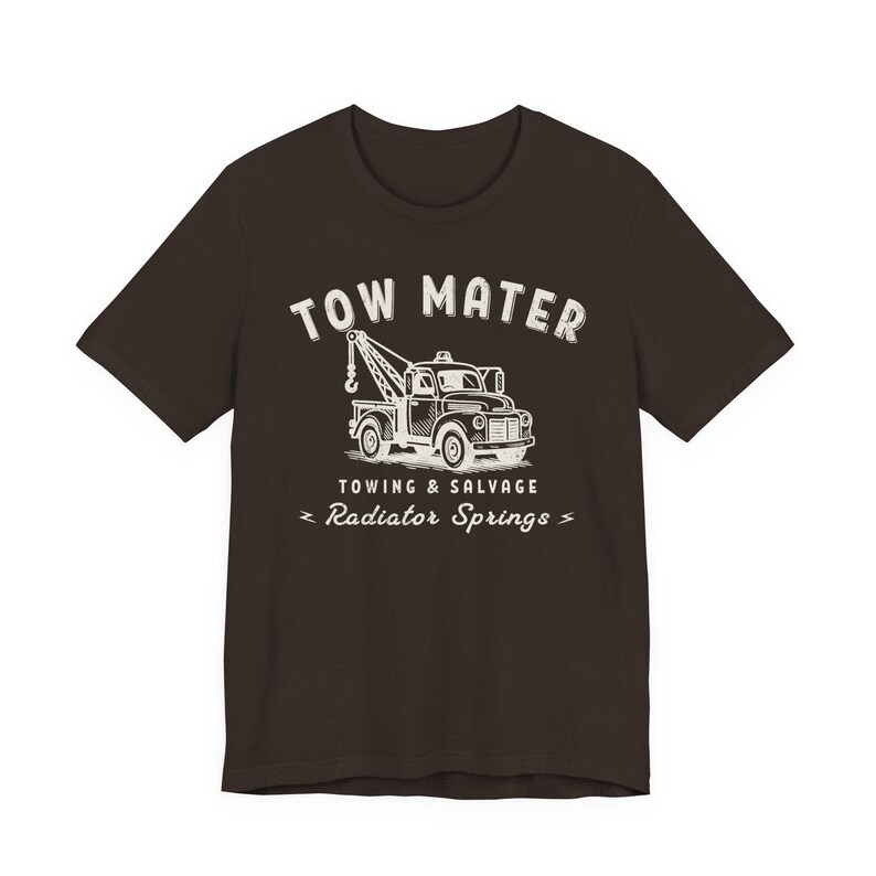 Pu&ograve; includere: T-shirt marrone scuro con una grafica bianca di un carro attrezzi e la scritta "TOW MATER" sopra. Sotto il camion ci sono le parole "TOWING & SALVAGE" e "Radiator Springs".